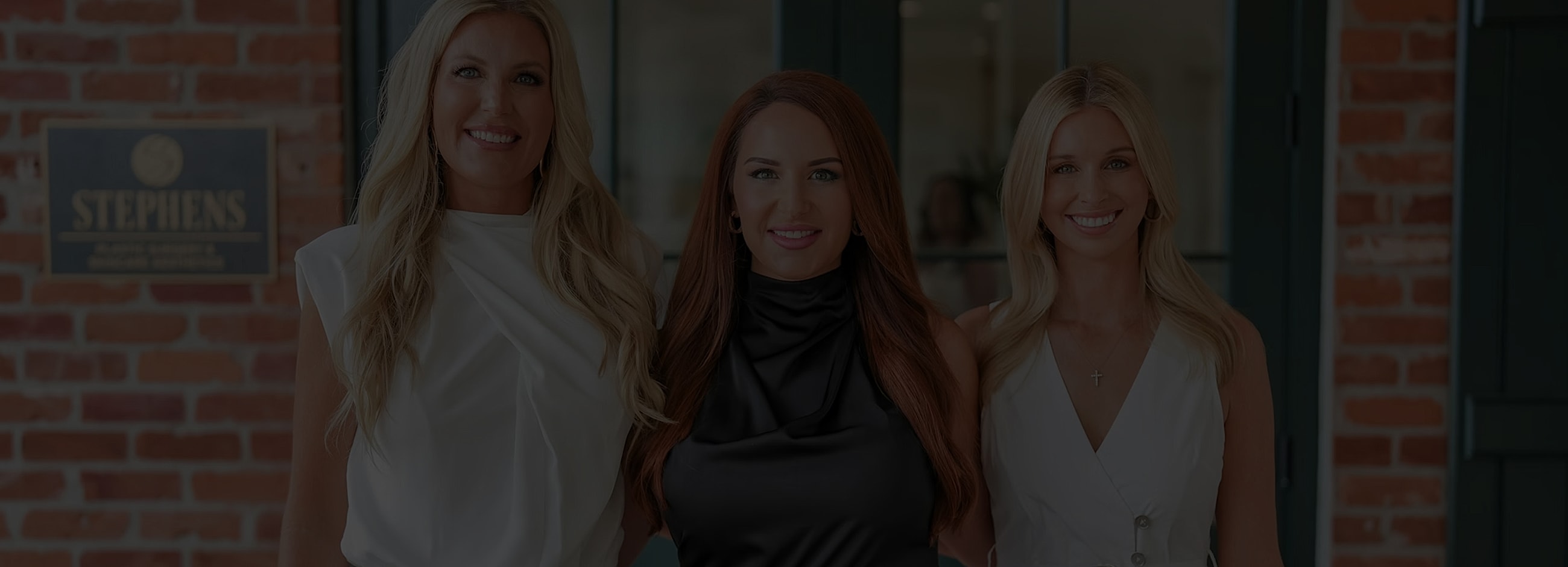 Three women smiling in front of a brick wall.